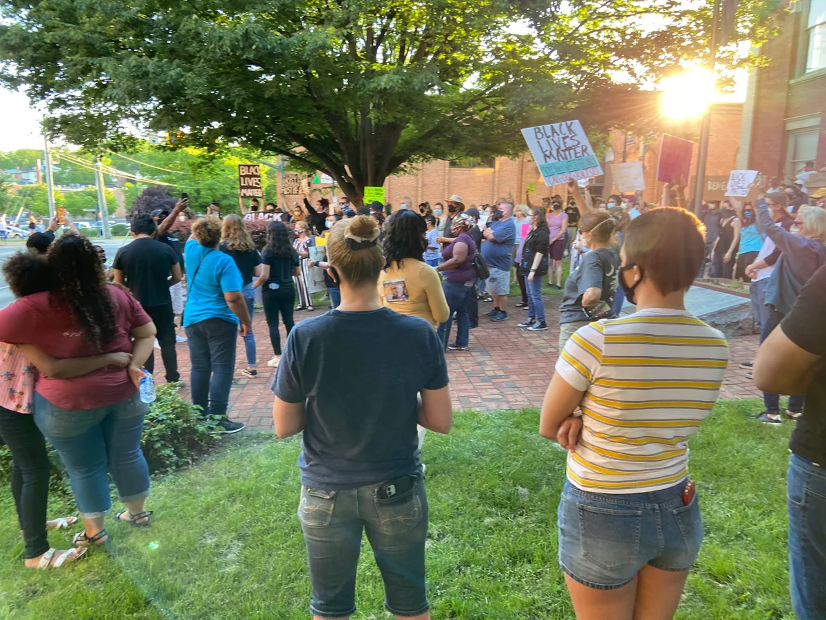 Crowd of masked protesters stands on a brick sidewalk and grass lawn beside historic brick buildings in warm golden-hour light, with a large leafy tree shading the scene. Handwritten signs reading “Black Lives Matter” rise above the gathering.