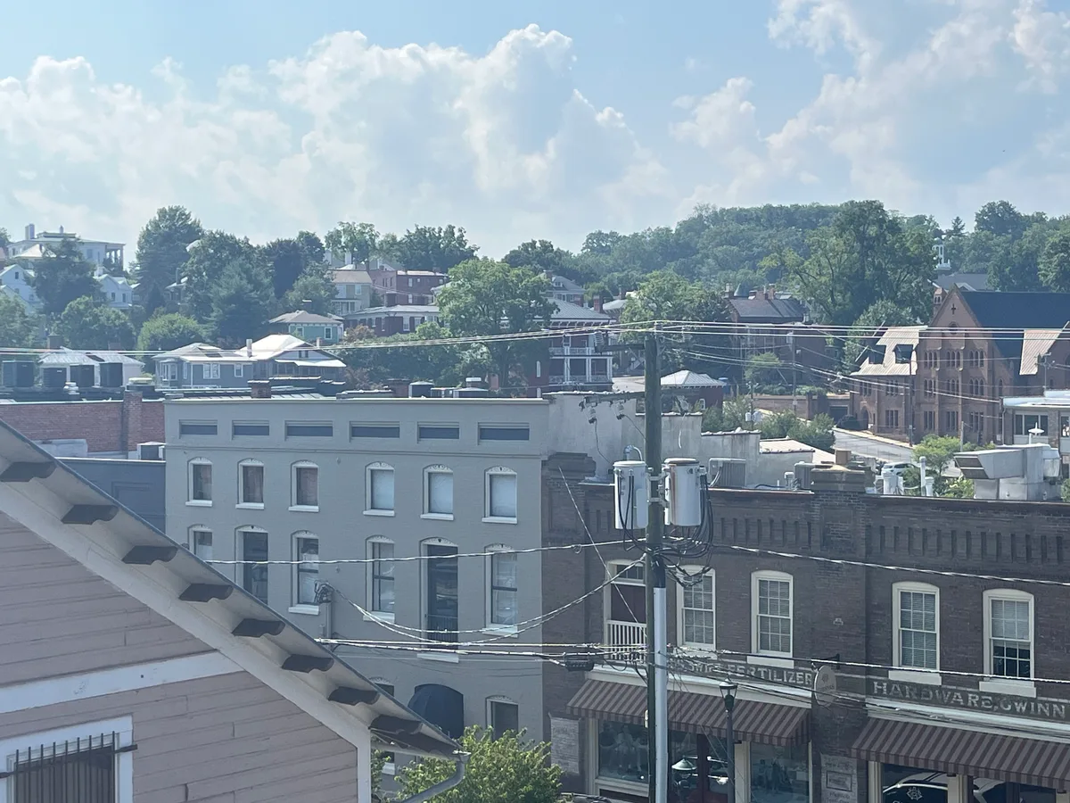 Sunlit view over Staunton’s historic downtown shows rows of Victorian brick and stucco buildings with tall sash windows, striped awnings, and old storefront signs beneath a web of utility wires. In the distance, leafy hills and tightly packed houses rise under a bright blue sky with towering clouds.