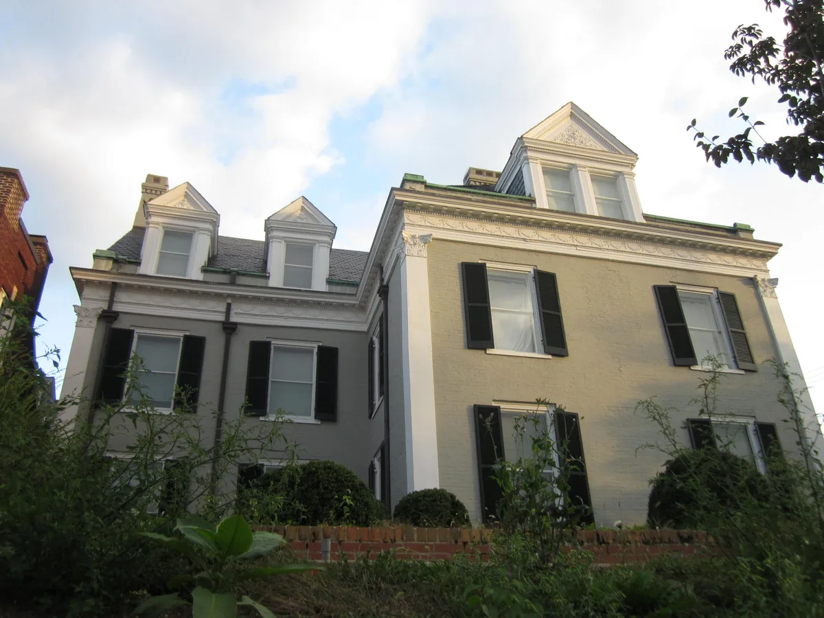 Low-angle view of two adjoining Victorian brick townhouses with ornate cornices, dormer windows, and black shutters, lit by soft golden-hour light. A low brick wall and overgrown greenery fill the foreground beneath a pale evening sky.