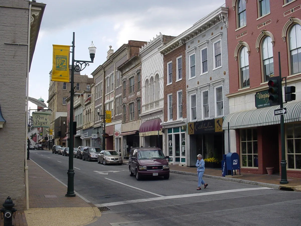 Victorian storefronts line a quiet downtown street in Staunton, with ornate brick and stucco facades, tall arched windows, striped awnings, and hanging shop signs along the block. Parked cars and a lone pedestrian cross beneath a soft overcast sky, emphasizing the intact historic streetscape.