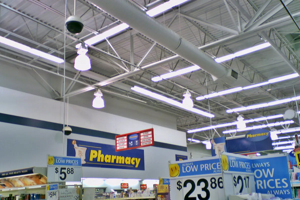 Bright fluorescent lights and exposed ceiling trusses hang over a Walmart pharmacy aisle, with large blue-and-yellow “Pharmacy” and “Always Low Prices” signs dominating the frame. The shot feels unfinished, with lighting fixtures and ductwork emphasized against the white interior walls.