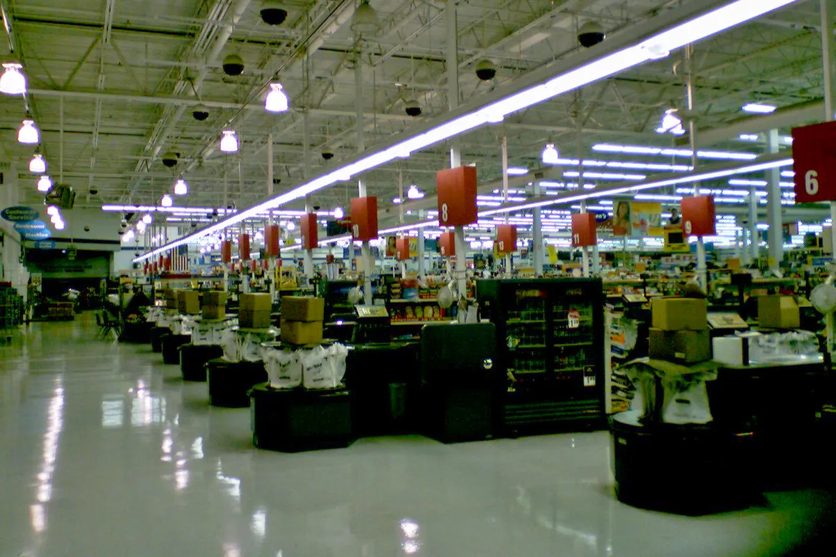 Wide view of a Walmart Supercenter interior with bright fluorescent ceiling lights, glossy white floors, and long rows of checkout lanes marked by red number signs. Boxes and stocked displays fill the foreground, while the open store stretches deep into the background under exposed industrial ceiling beams.