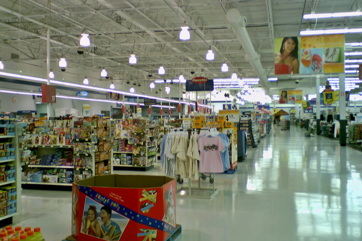 Brightly lit Walmart interior with rows of aisles, clothing racks, and stacked merchandise beneath a high industrial ceiling. The glossy floor reflects the overhead lights, and colorful promotional signs hang above the wide open sales area.