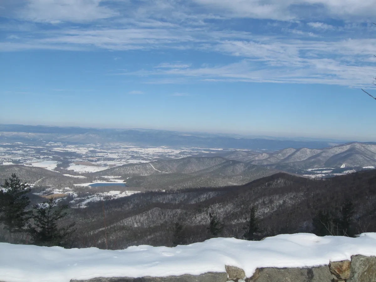 Snow blankets the stone ledge at the overlook while layered blue ridges and a broad valley stretch into the distance under a pale winter sky. Patchy white fields and dark, forested hills create a wide panoramic view from the summit.