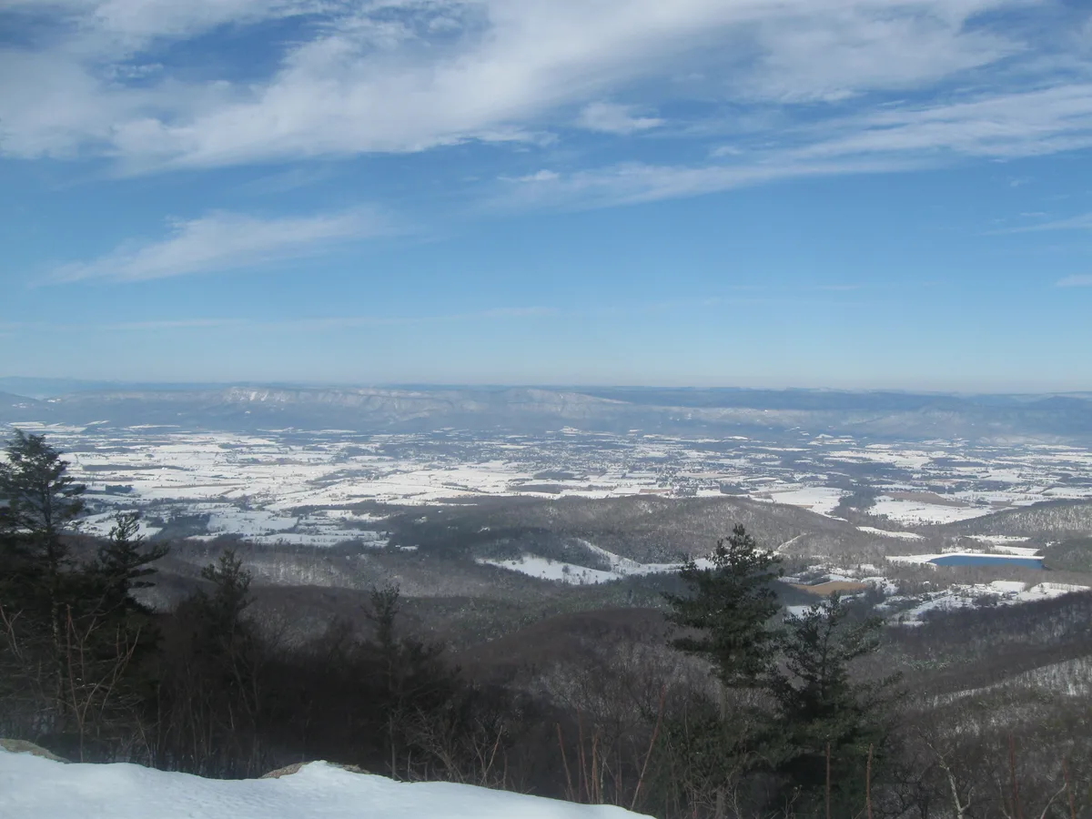Snow-covered ridges and dark evergreen trees frame a wide view across the Shenandoah Valley, with patchwork white fields stretching to distant blue mountains under a pale sky. The overlook is taken in clear winter light, with wispy clouds overhead and a bright strip of snow along the cliff edge in the foreground.