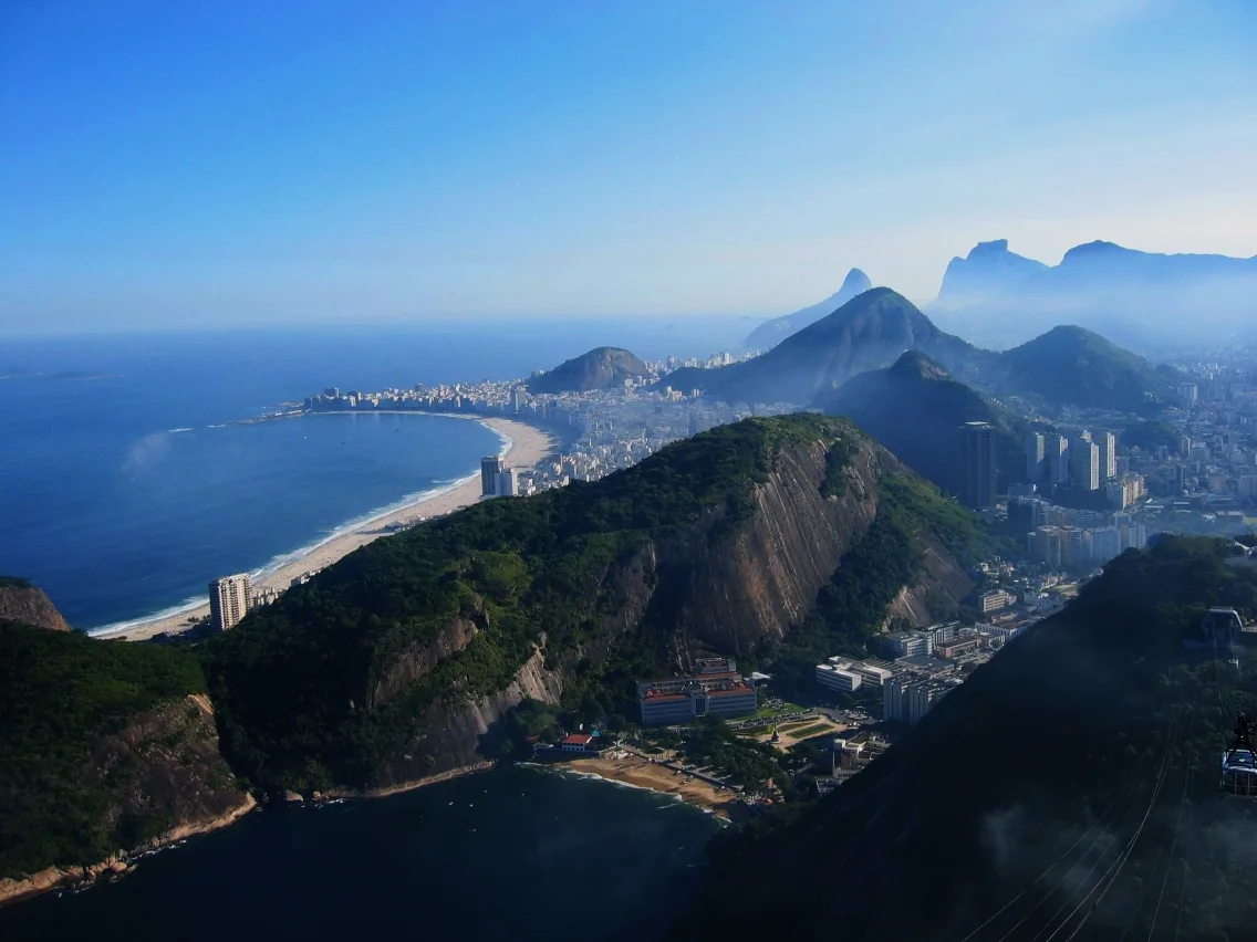 Wide panoramic view from a mountain summit over Rio de Janeiro, with sweeping blue bay, crescent beaches, and dense city neighborhoods tucked between steep green granite peaks. Clear golden light and light haze soften the distant mountains and coastline.