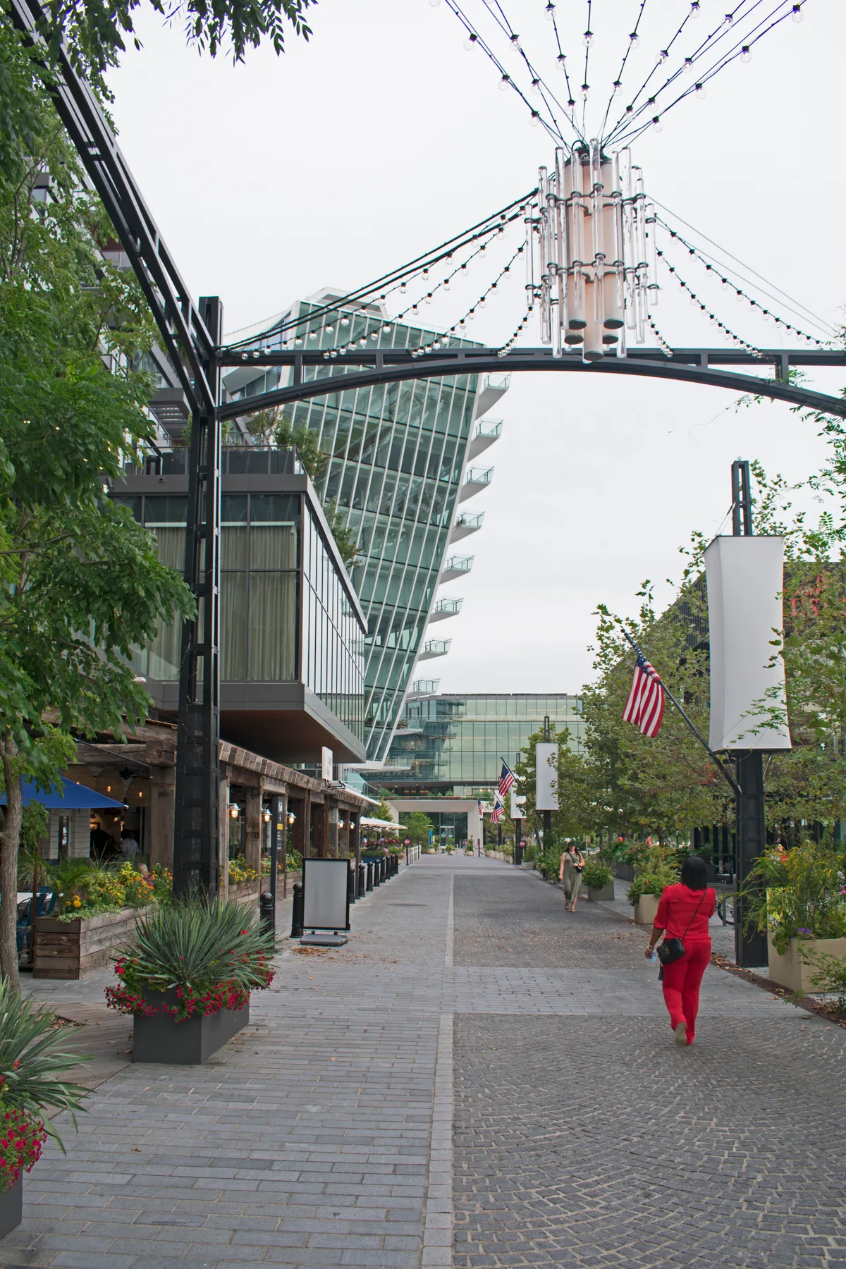 A cobblestone pedestrian street at The Wharf is lined with planters, string lights, and hanging fixtures, leading toward sleek glass buildings. A few people walk beneath American flags, with a striking slanted tower rising in the background under an overcast sky.