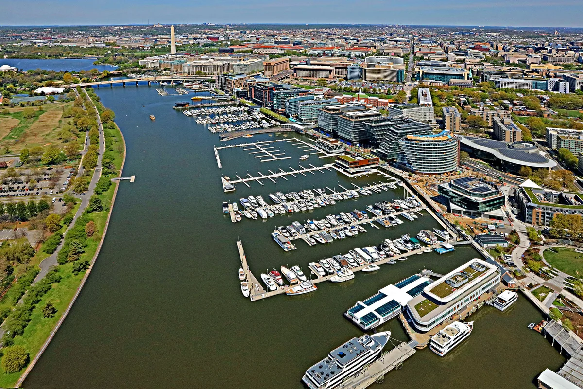 Wide aerial view of The Wharf’s marina stretching along the Anacostia, with rows of docked boats, curved waterfront buildings, and the Washington Monument rising in the distance. The water forms a dark central corridor bordered by green shoreline on the left and dense city blocks on the right.