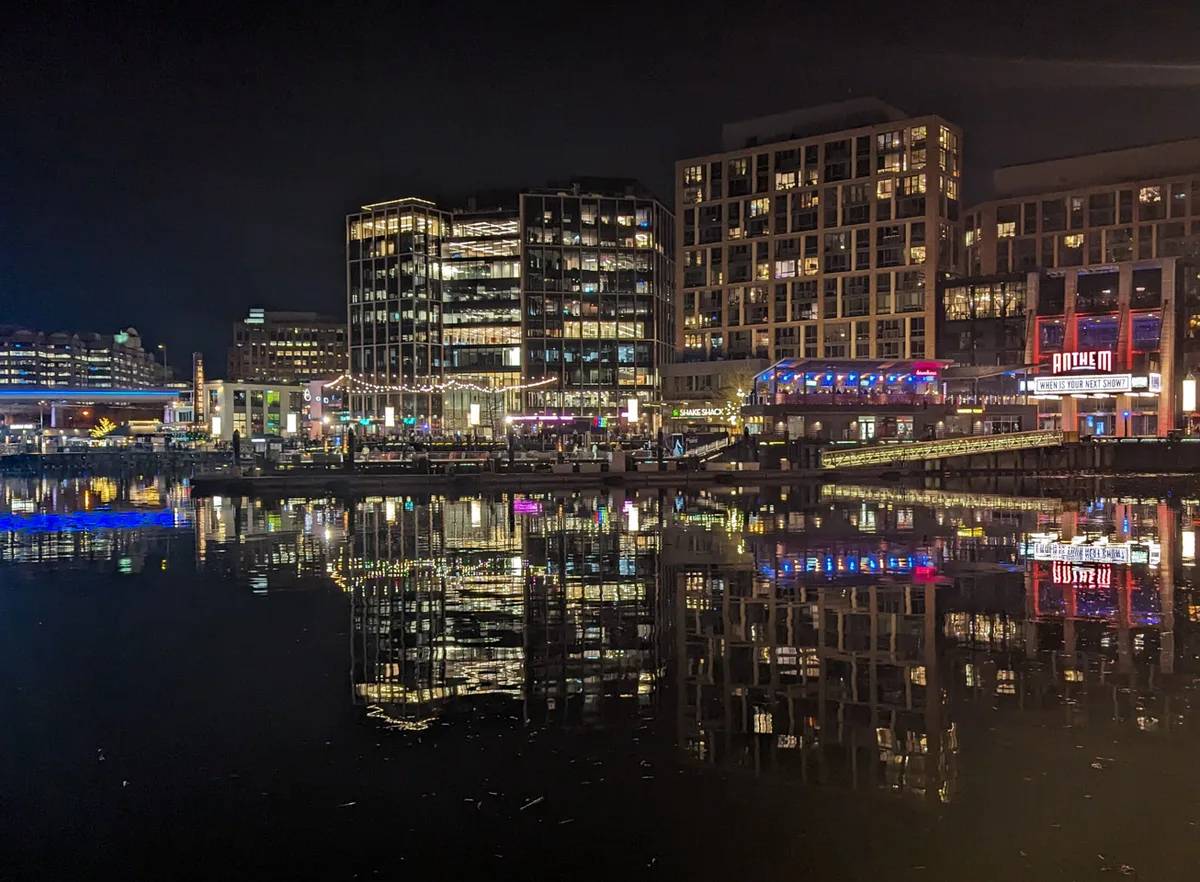 Nighttime at The Wharf shows lit modern apartment and entertainment buildings along the waterfront, including the glowing Anthem sign, reflected in the calm dark water. Blue, gold, and white lights shimmer across the marina-like surface under a black sky.