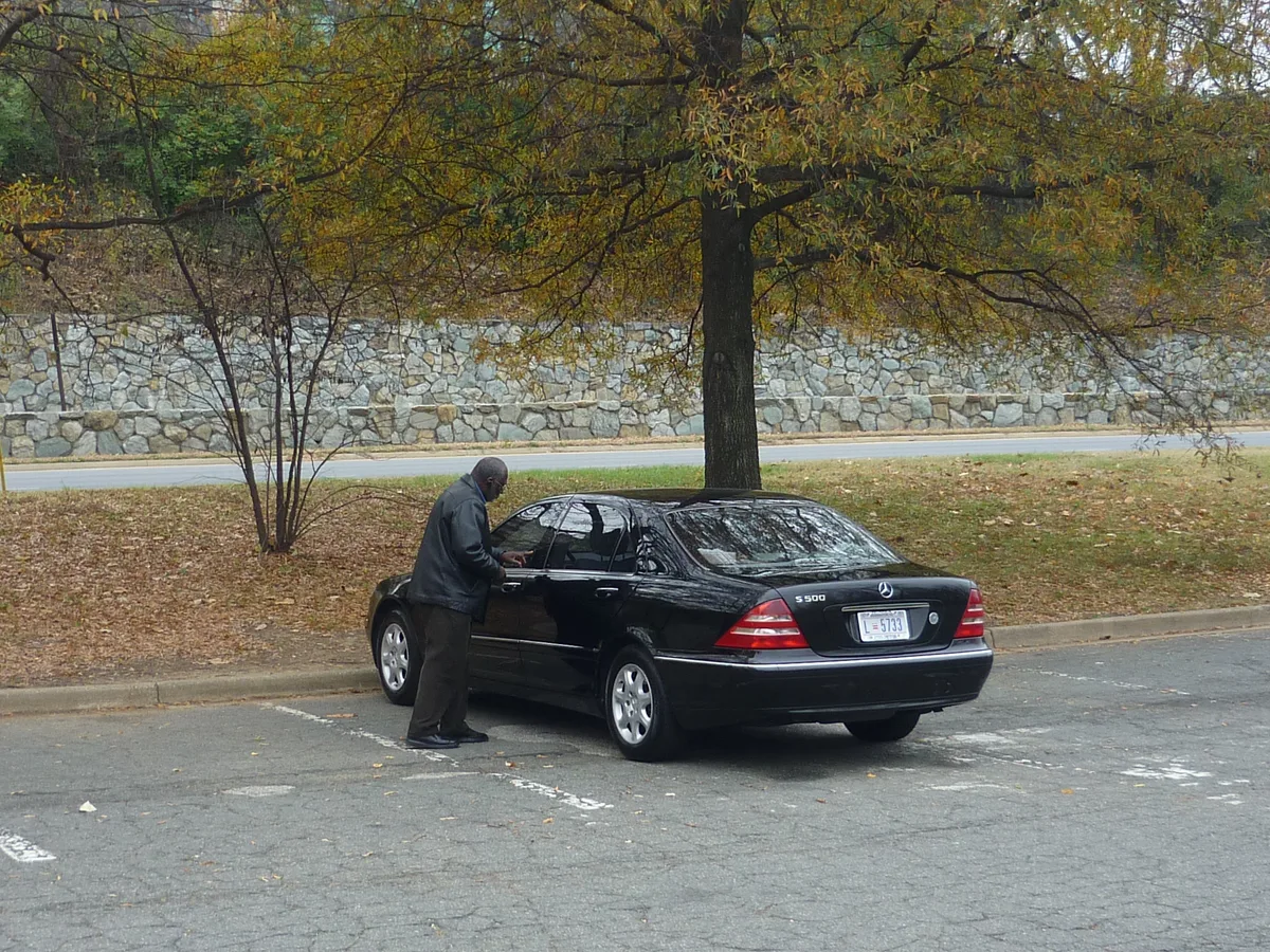 A black Mercedes sedan is parked in a nearly empty lot beneath a tall autumn tree, with a man standing by the driver’s door. A stone retaining wall and quiet road run across the background under muted fall light.