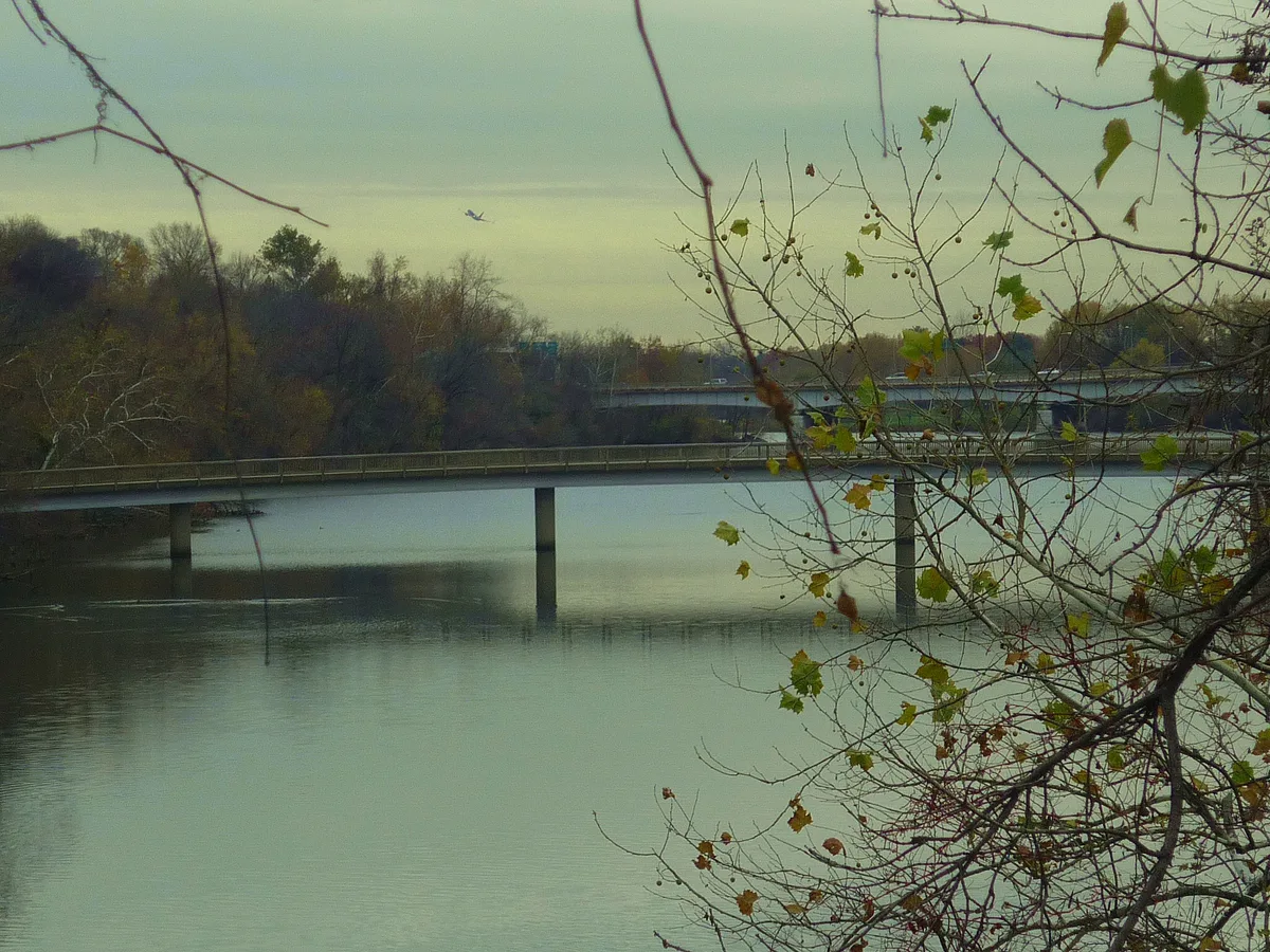 A low footbridge stretches across the calm Potomac, framed by bare winter branches and a few lingering leaves in the foreground. Soft golden light and a hazy sky give the river and distant tree line a quiet, muted look.