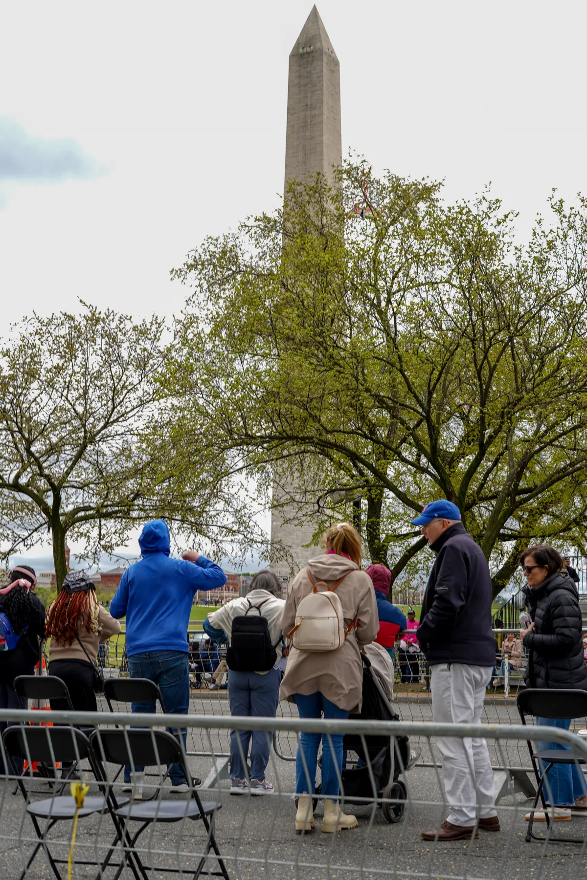 Spectators in jackets and hats stand behind metal barricades and folding chairs, looking toward the parade route with the Washington Monument rising above the spring-green trees. The overcast sky softens the scene and leaves the tall stone obelisk centered in the background.