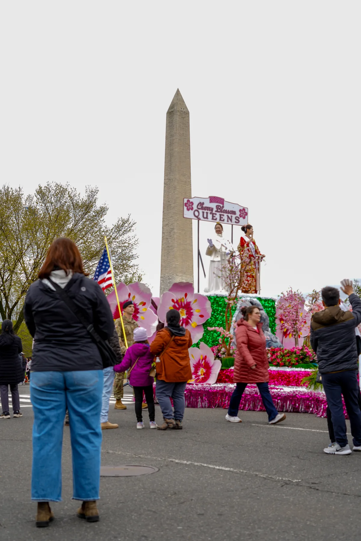 A cherry blossom parade float passes the Washington Monument, topped with a “Cherry Blossom Queens” sign and covered in oversized pink flower decorations. Spectators in jackets watch from the street beneath an overcast spring sky.