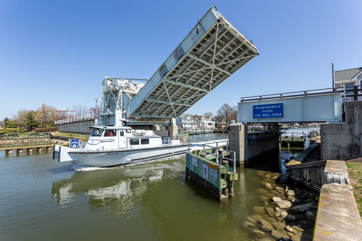 A white workboat passes beneath the raised Knapps Narrows drawbridge, with the steel span lifted high over the green channel. Sunny blue sky, waterfront houses, and dock pilings frame the narrow Chesapeake waterway.