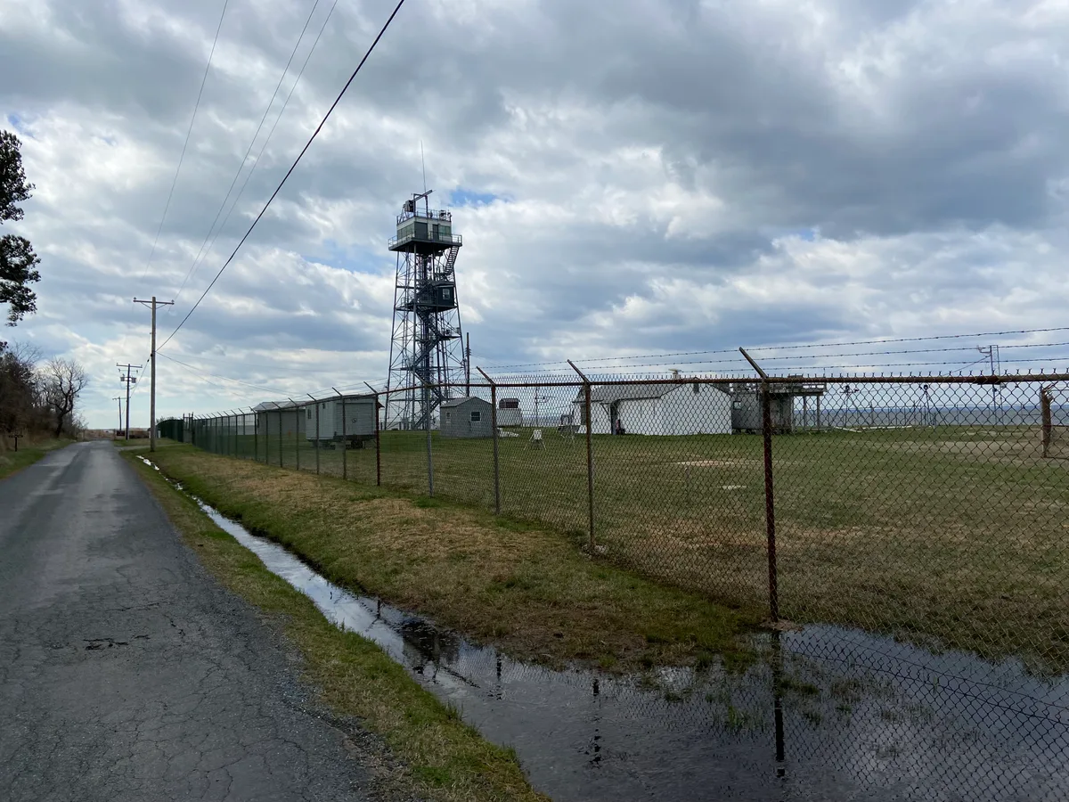A narrow rural road runs beside a chain-link fence and drainage ditch toward a tall metal tower and low utility buildings on flat marshland. Heavy gray clouds hang overhead, with the bay visible in the distance beyond the fenced site.