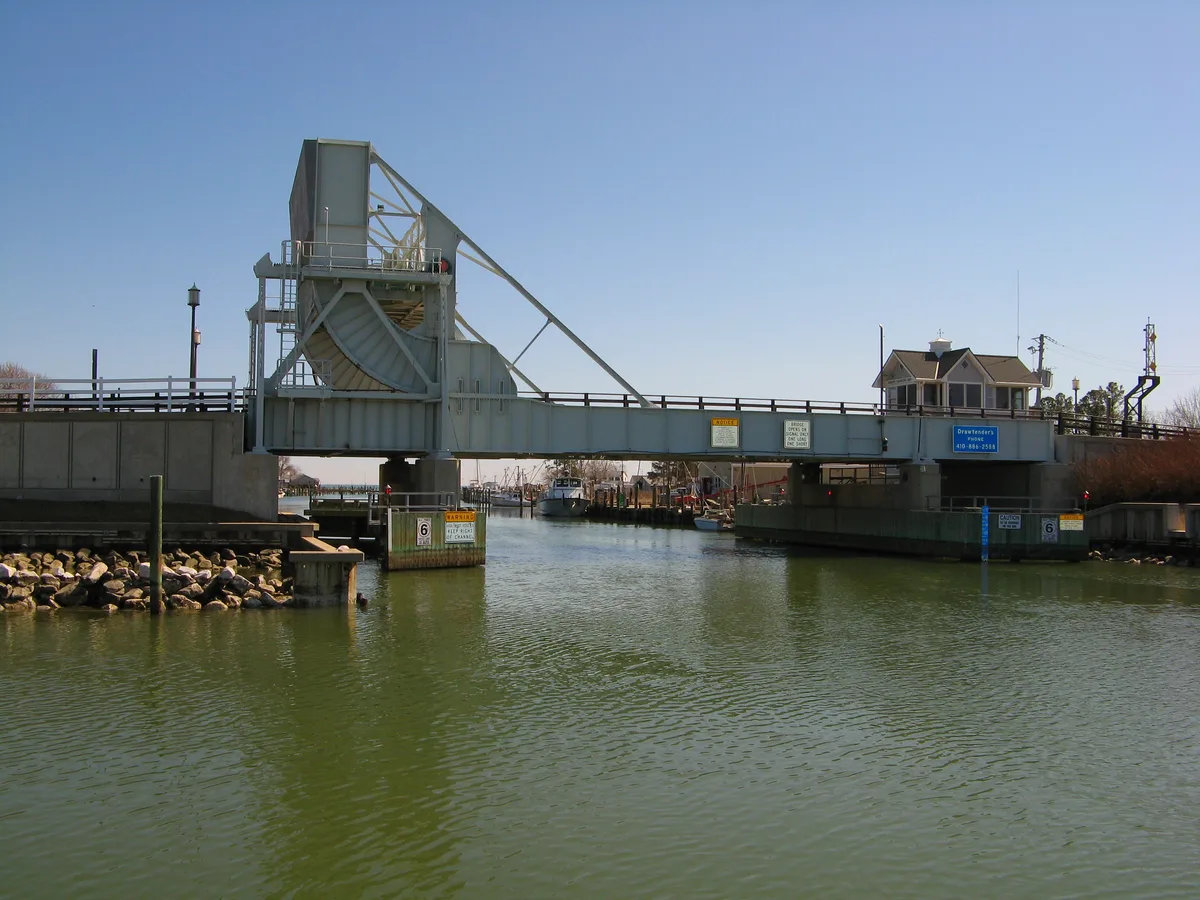 Maryland Route 33 drawbridge spans a narrow Chesapeake channel, its steel lift tower raised against a clear blue sky. Green water fills the foreground, with harbor boats and Tilghman Island waterfront buildings visible beyond the bridge.
