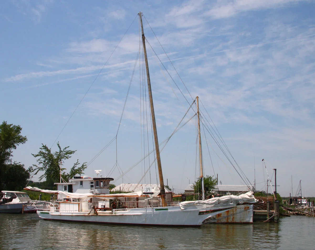 A white Chesapeake skipjack with tall wooden masts is tied up along a quiet Tilghman Island dock, its sails furled and rigging crossing against a pale blue sky. Working boats, a low waterfront shed, and calm water frame the weathered maritime scene.