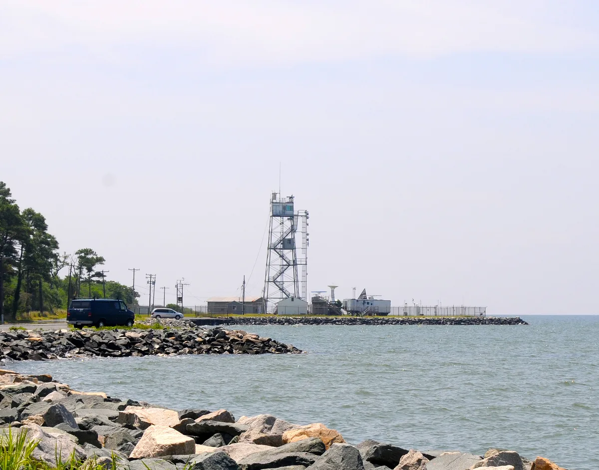 A tall steel radio tower rises from a low industrial compound on the edge of Tilghman Island, with a rocky breakwater and choppy gray-blue water in the foreground. Sparse pine trees and a couple of parked vehicles line the shore under a bright, hazy sky.