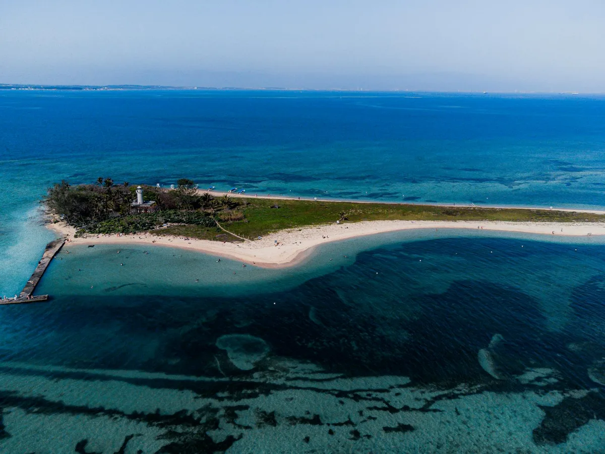 A high, wide aerial view shows a tiny sandy island ringed by clear turquoise water and darker blue channels, with a white lighthouse near the tree line and a narrow dock extending into the bay. Small boats and tiny figures dot the shoreline beneath a pale, hazy sky.