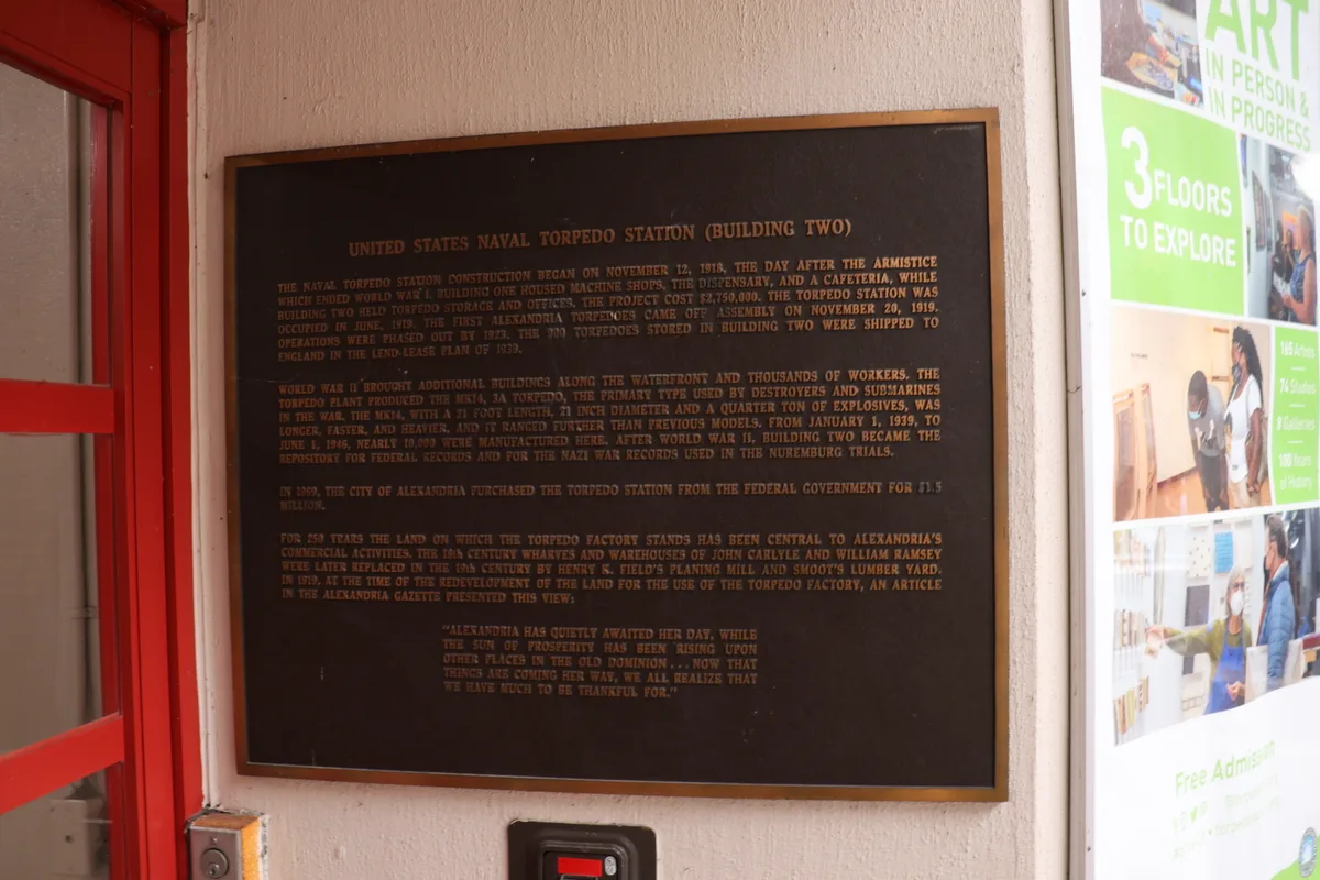 A dark bronze historical plaque mounted on a cream wall marks the United States Naval Torpedo Station, framed by a bright red industrial doorway on the left. To the right, a colorful poster for the Torpedo Factory peeks in, contrasting the building’s wartime past with its present-day art center.