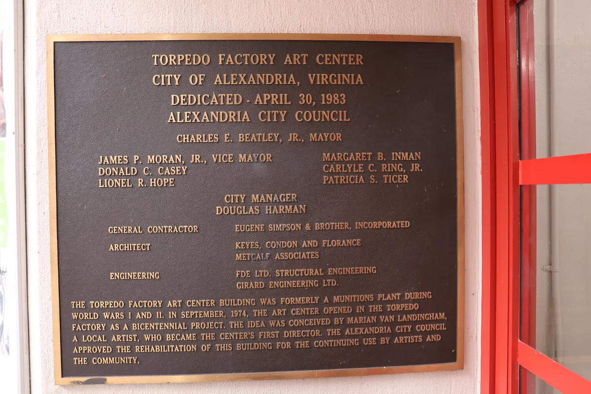 A bronze dedication plaque is mounted on a pale interior wall, with raised lettering that honors the Torpedo Factory Art Center’s 1983 opening in Alexandria. A bright red doorframe or railing cuts along the right edge, adding a vivid industrial accent to the close-up composition.