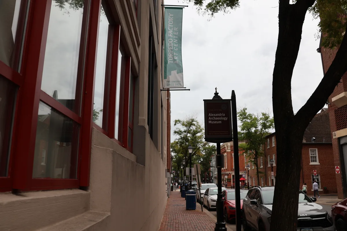 Red brick Torpedo Factory Art Center walls with tall red-framed windows line the left side of a quiet Alexandria sidewalk, where a vertical banner hangs above the street. A sign for the Alexandria Archaeology Museum, parked cars, and leafy trees under a bright overcast sky add to the downtown scene.