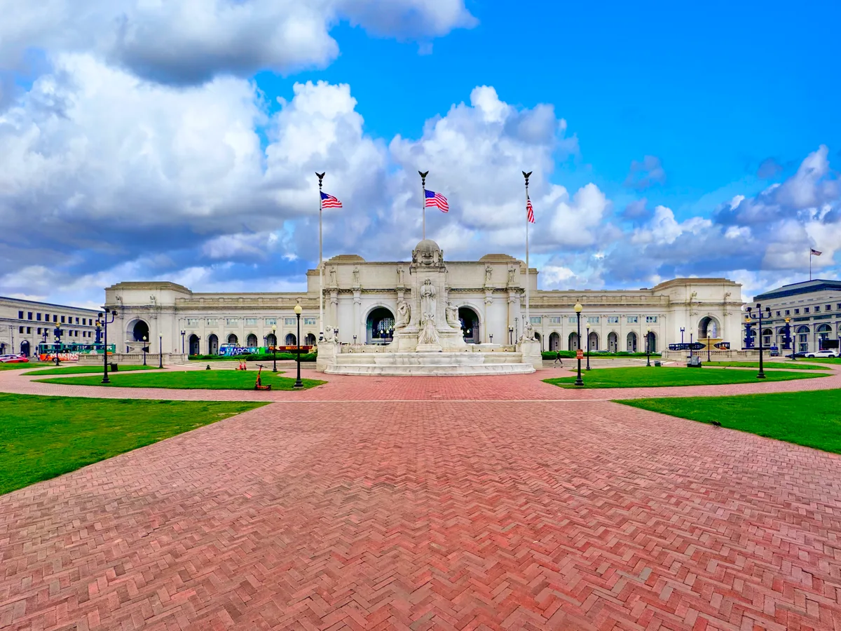 Union Station’s grand Beaux-Arts façade stretches across the frame behind a central marble fountain, with three American flags flying above the entrance under a bright, cloud-filled sky. A red brick plaza and manicured green lawn lead the eye toward the symmetrical arches and colonnades of the station.