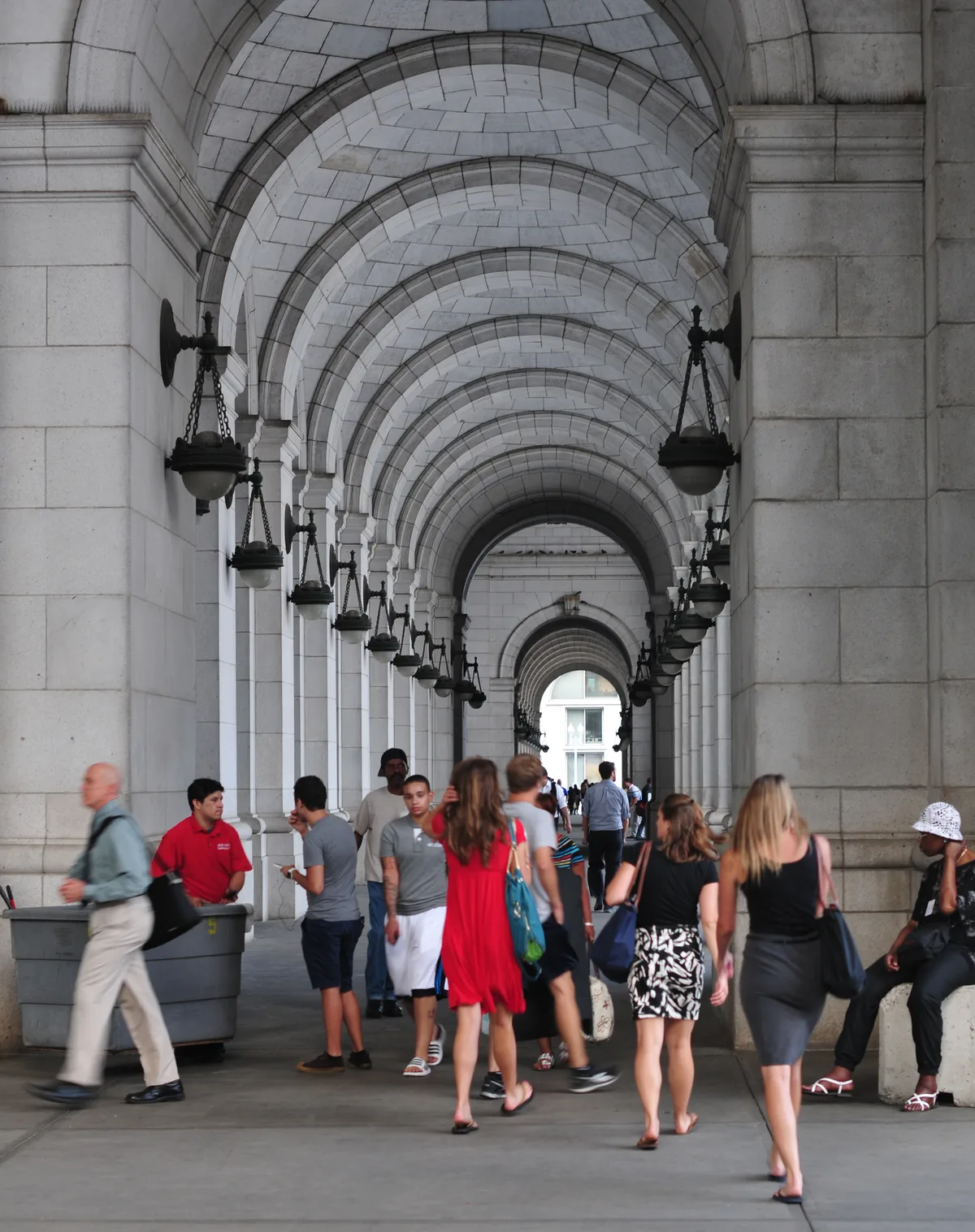 Union Station’s grand stone arcade stretches into the distance under a series of repeating barrel vault arches, with hanging black lanterns lining both sides. People walk through the shaded concourse beneath the pale Beaux-Arts ceiling, while bright daylight shows at the far end.