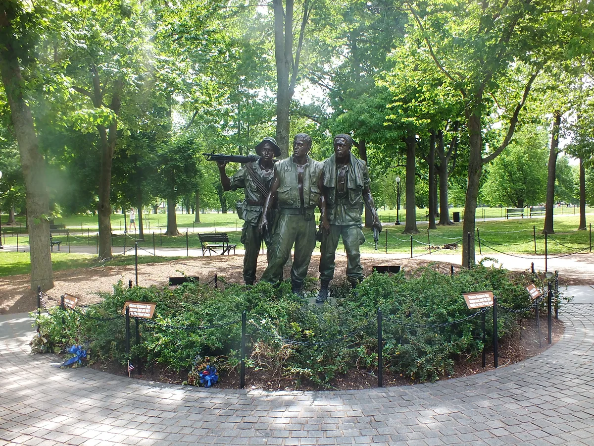 Three bronze soldiers stand in a small circular grove, surrounded by low shrubs, black chain fencing, and curved brick paving under tall green trees. Sunlight filters through the leaves, giving the memorial a quiet, dappled look.