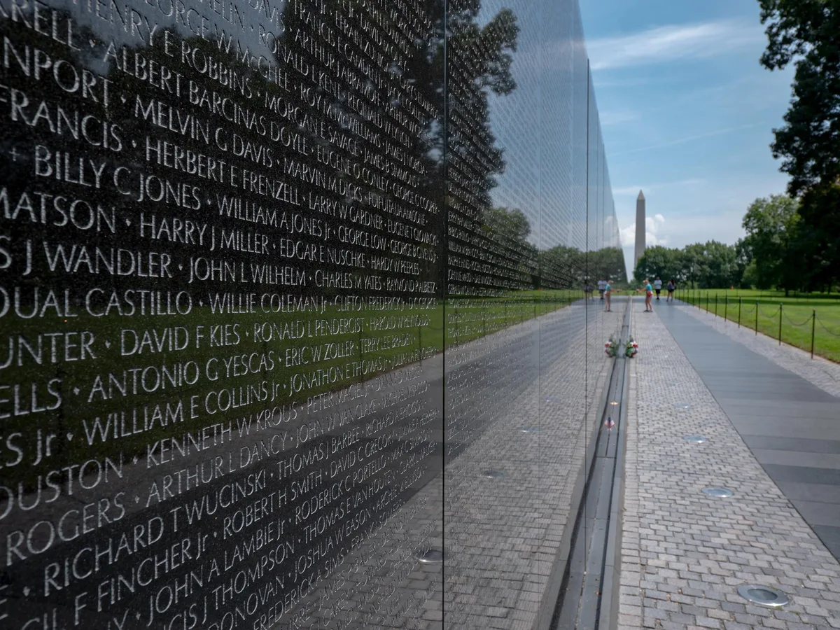 The black granite Vietnam Veterans Memorial wall fills the left side with engraved names, its polished surface reflecting the blue sky, trees, and walkway. In the distance, the Washington Monument rises beyond a few visitors and flower tributes along the base of the wall.