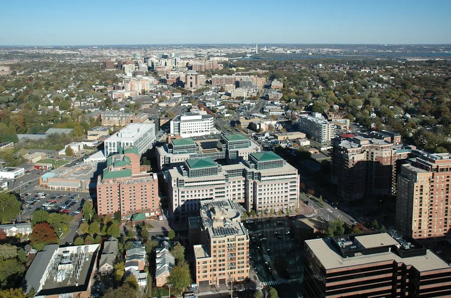Wide daytime view over Arlington and toward Georgetown, with clusters of brick and glass buildings in the foreground, tree-filled neighborhoods stretching across the middle distance, and the DC skyline faintly visible on the horizon. The scene is crisp and sunlit, with long shadows cutting across the streets and rooftops.