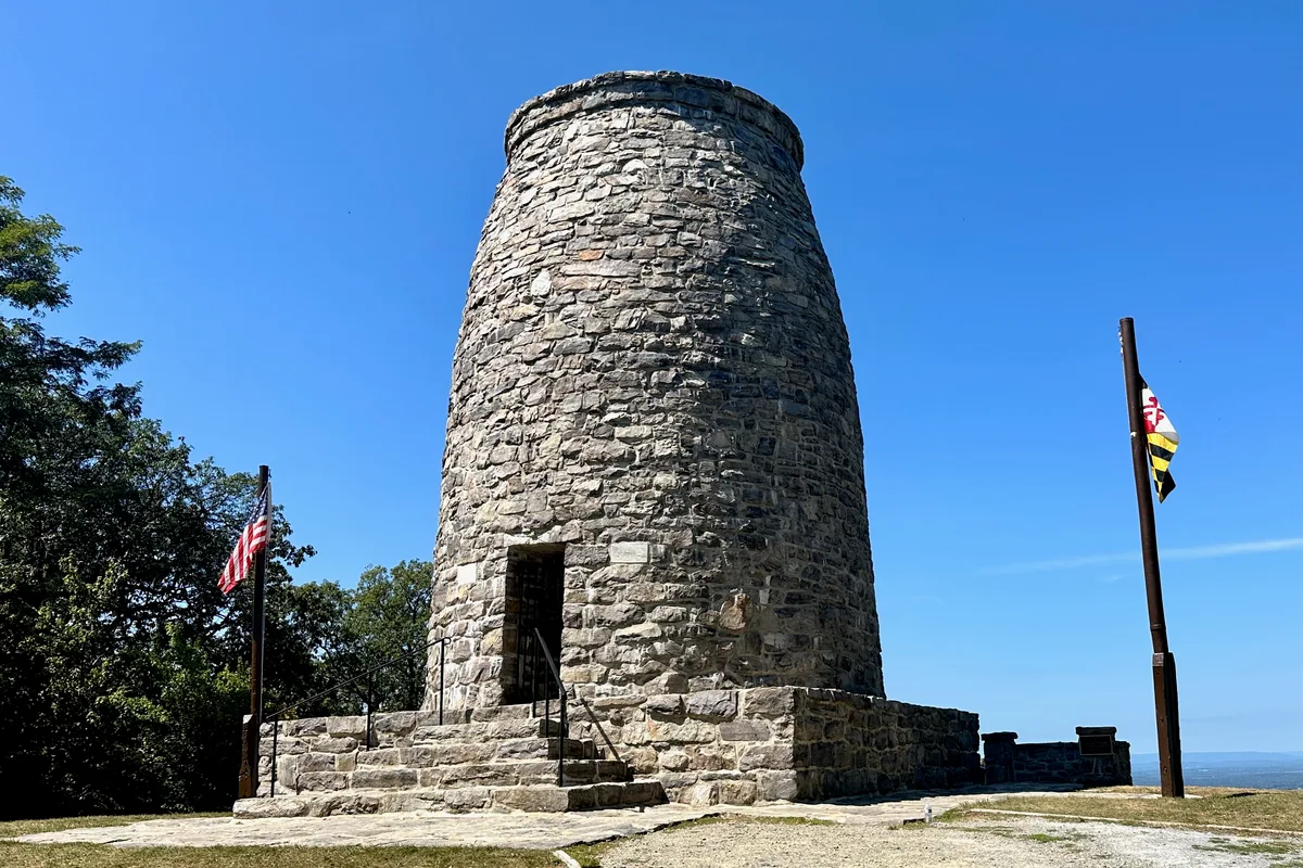A round stone observation tower rises from a rocky platform, framed by American and Maryland flags against a clear blue sky. Trees crowd the left side while the open overlook and distant horizon stretch out on the right.