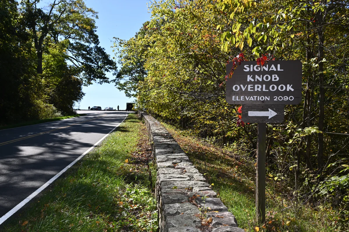 A roadside overlook sign reading “Signal Knob Overlook” stands beside a stone retaining wall and a two-lane mountain road under bright midday sun. Trees with early fall color frame the view, and the road rises toward a small pullout against a clear blue sky.