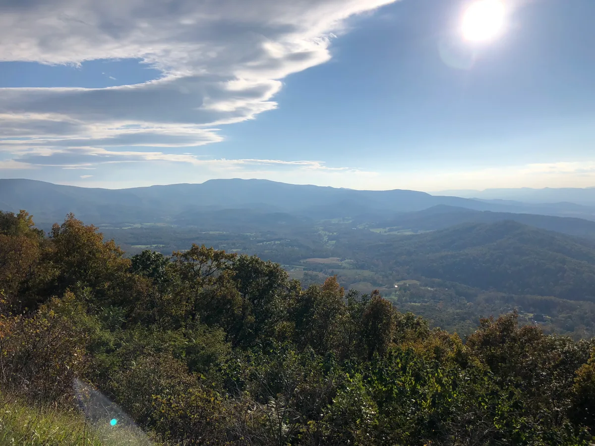 Sunlight hangs high over a broad Shenandoah Valley panorama, with layered blue ridges fading into the distance beneath a bright sky streaked with thin clouds. Dense green foreground trees frame the overlook, and pale fields and a faint river curve are visible in the valley below.