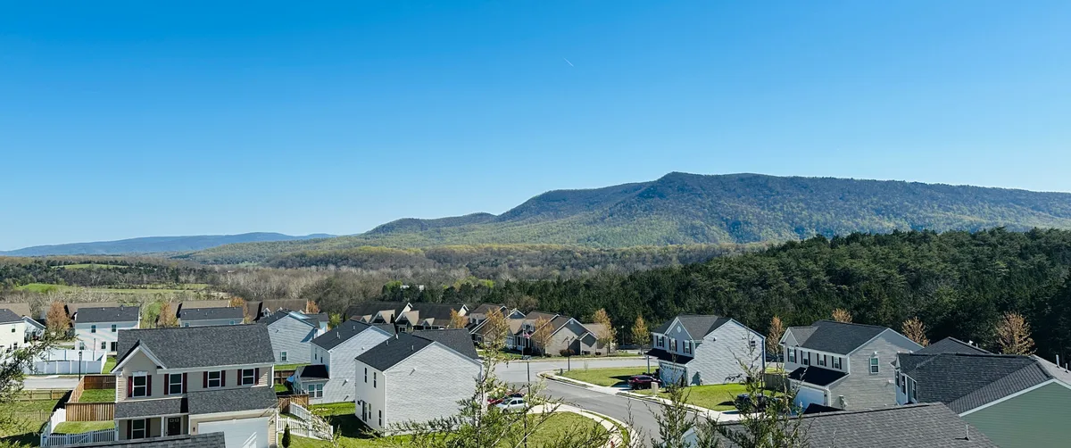 Wide view over a suburban neighborhood with rows of gray-roofed houses in the foreground, backed by a dense green tree line and the broad blue ridge of Massanutten Mountain under a clear sky. The scene is lit by bright late-day sun, with the valley stretching out in soft layers toward the horizon.