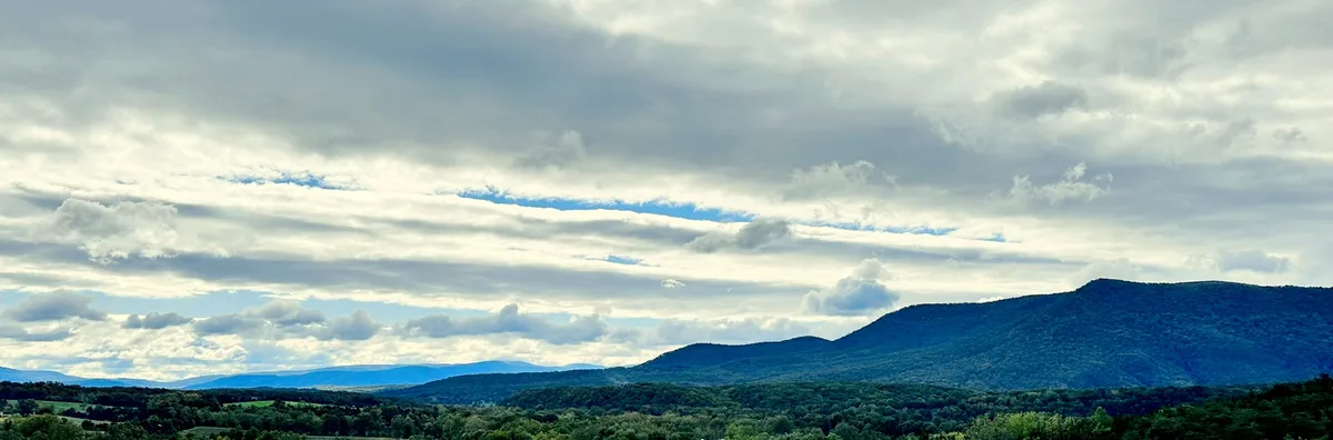 Wide panoramic view over the Shenandoah Valley from Woodstock Tower, with a broad green foreground and a long blue-green mountain ridge stretching across the horizon. A bright, cloud-filled sky hangs over the landscape, with soft golden light and a faint river bend visible in the distance.