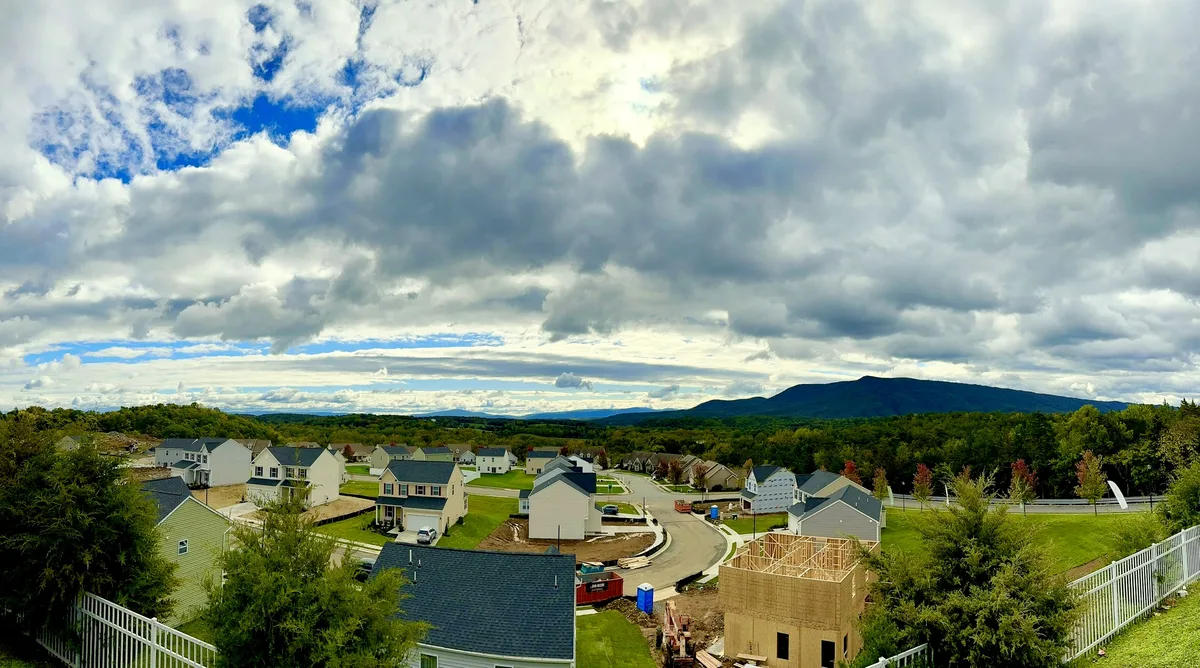Wide panoramic view from the tower over a suburban neighborhood spreading across the Shenandoah Valley, with rows of new houses, curving streets, and a forested mountain ridge in the distance. Thick clouds hang over the scene, breaking to reveal patches of blue sky above the green landscape.