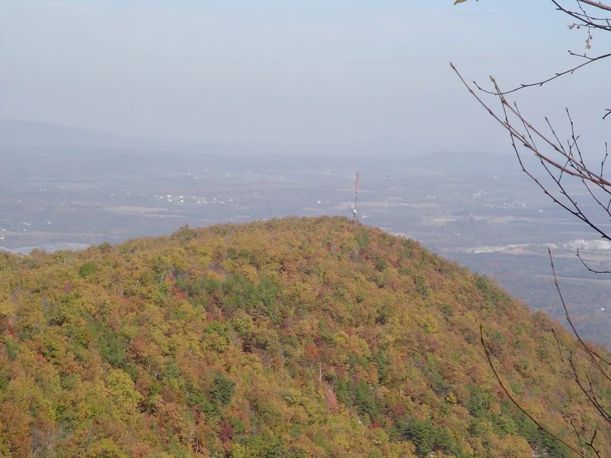 A broad autumn view from Woodstock Tower looks out over a forested ridge covered in patchy fall color, with a small communications mast on the summit. Hazy Shenandoah Valley layers stretch into the distance under a pale sky, with bare branches framing the right edge.