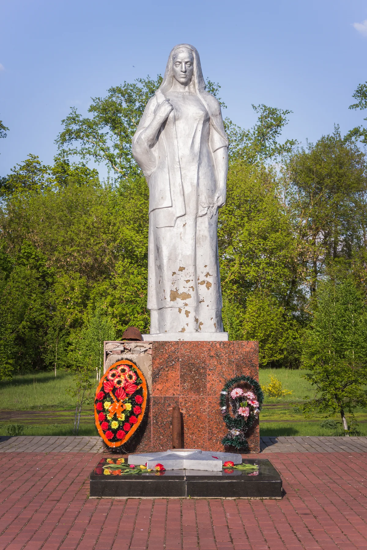 A tall white statue of a robed woman stands on a red granite base, with colorful wreaths and scattered flowers at its foot. It’s set on a brick-paved platform against a backdrop of green trees and bright blue sky in daylight.