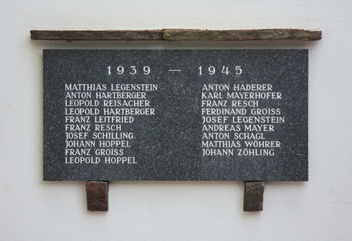 A dark granite memorial plaque is mounted on a white wall, with the years “1939 — 1945” engraved above two columns of white names. Rough stone and metal brackets frame the plaque, giving it a simple, austere look.