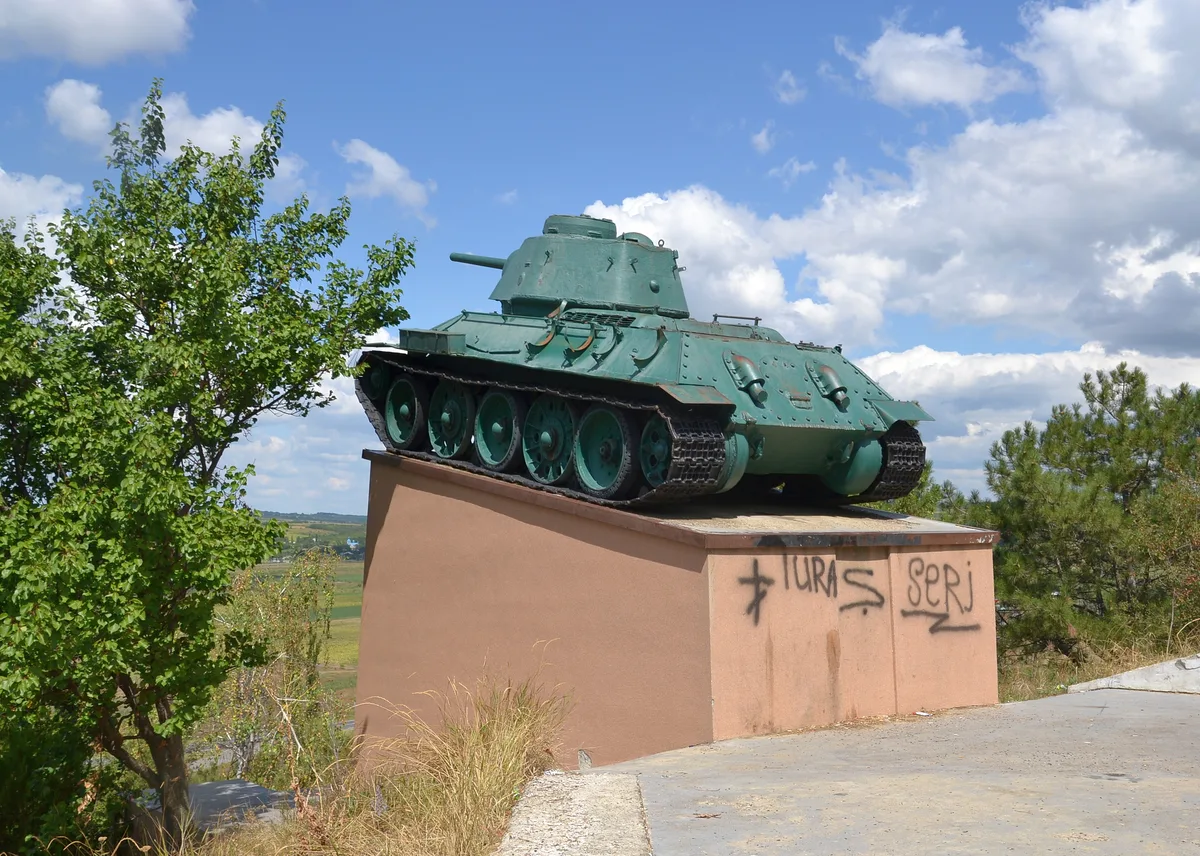 A green World War II-era tank sits on a tan pedestal beside a paved overlook, with graffiti on the base and trees framing the scene. Bright midday light and a blue sky with scattered clouds open out over the surrounding countryside.