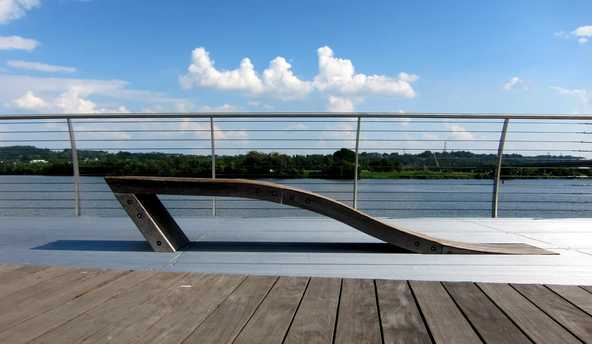 A sculptural curved wooden bench sits on the waterfront deck at Yards Park, framed by a cable railing and the wide Anacostia River beyond. Bright blue sky and scattered clouds give the scene a clean, open summer feel.
