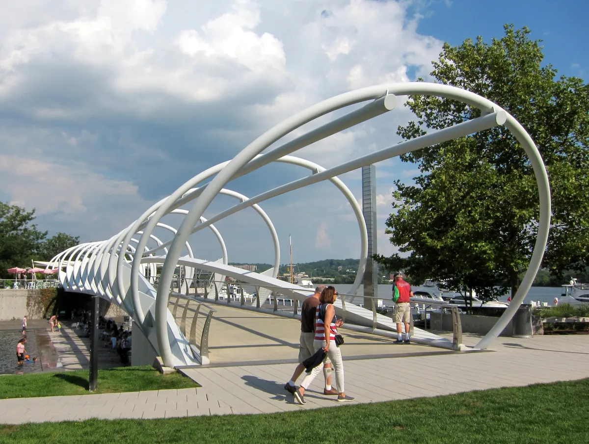 Curving white steel arches form a sculptural pedestrian bridge over Yards Park, with people walking across the broad deck beside the Anacostia River. Blue sky and billowing clouds frame the modern waterfront scene, with trees, boats, and a shallow water feature below.