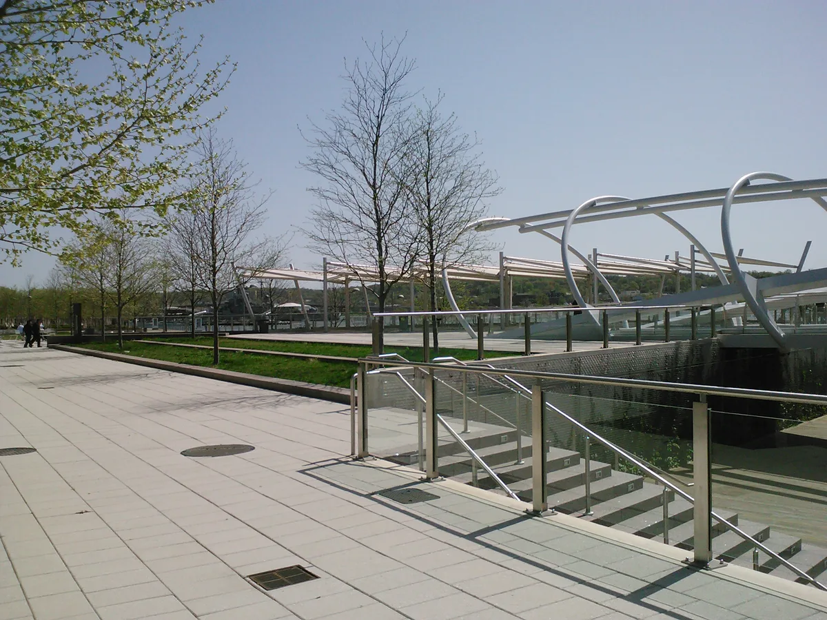 Wide view of Yards Park’s clean, modern waterfront plaza, with pale paving, glass railings, and a sculptural white cable bridge crossing over a sunken water feature. Leafing trees and a clear sky frame the open urban landscape.